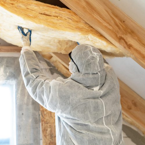 Back view of one foreman in overalls working with rockwool insulation material, fasten warmth layer on ceiling and wall, standing inside new house under construction